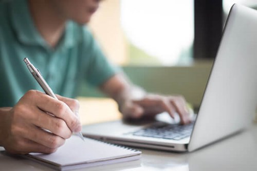 person typing on a laptop and writing notes in notepad 